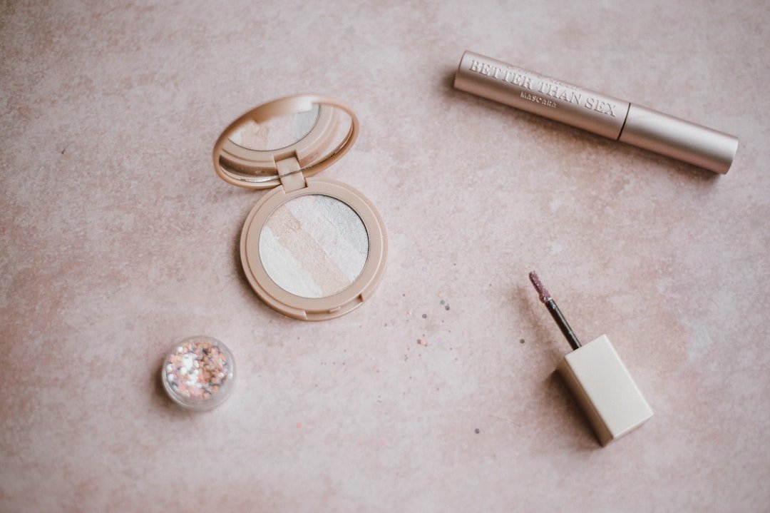 Flatlay of cosmetics products including powder, highlighter, mascara, and lip gloss arranged on a neutral surface