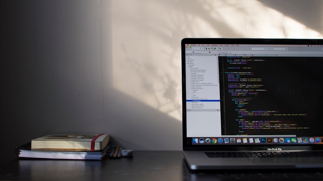 MacBook Pro on a dark desk showing a code editor with stacked books beside it and sunlight on the wall behind