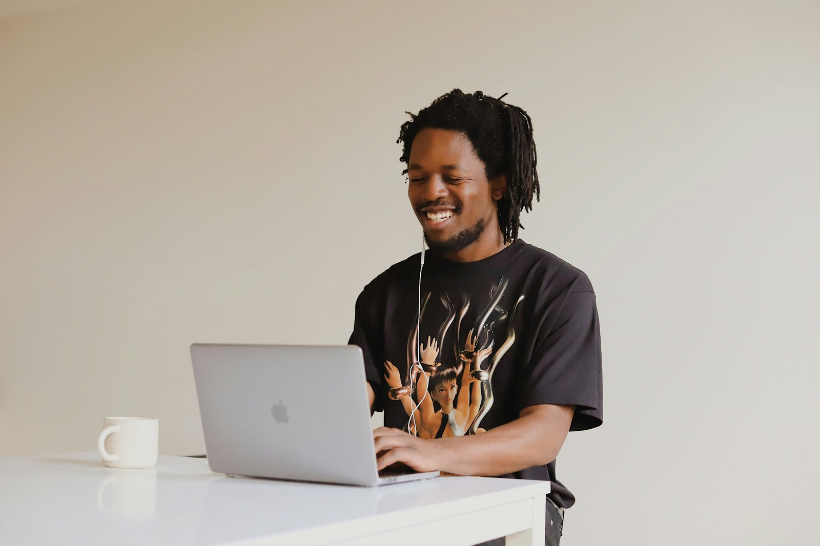 Smiling man with dreadlocks wearing earbuds and a black graphic t-shirt typing on a MacBook at a white desk