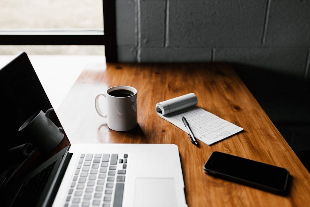 MacBook laptop, white mug, handwritten notepad, and smartphone on a warm wooden desk near a window